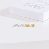 Front view of Pusheen Lazy Stud Earrings, which are unmatched poses of Pusheen the Cat lounging. The earrings on the left are sterling silver and on the right are the gold vermeil earrings. Behind the earrings sits the white packaging case that the earrings come in.