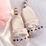 Pusheen Plush Slippers lying on a white fluffy surface next to a light pink pillow. Plush slippers have Pusheen's face embroidered accompanied by ears that extend off the toe box. Off the back of the slippers are Pusheen's striped tails. 