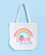 Pusheen Rainbow Tote Bag lies on a light blue surface. The white canvas tote bag features a grey Pusheen the Cat surrounded by a pink, yellow, and blue rainbow and white clouds. Pusheen sits atop the pink and blue colored phrase “Love Yourself.” 