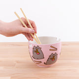A hand holding a pair of plastic chopsticks into a pink bowl featuring a repeating pattern of chop sticks and Pusheen eating a bowl of ramen. The bowl has two holes around the edges, one square hole on one edge, and a square indent on the rim of the bowl on the opposite side. 