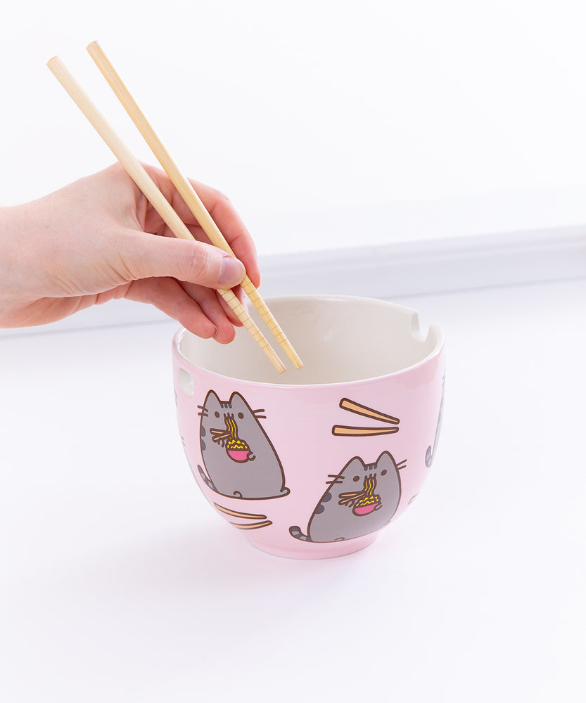 A hand holding the chopsticks over the Pusheen Ramen Bowl from the left, in front of a white background. 