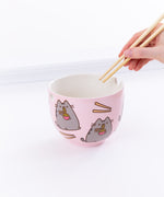A hand holding the chopsticks over the Pusheen Ramen Bowl from the right, in front of a white background. 