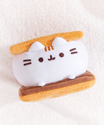 S’mores-inspired plush laying down on a white fluffy backdrop. The Squisheen marshmallow element is white, the graham cracker plush are light brown, and the chocolate slab is a dark brown color.