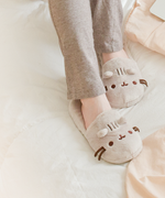 Model wearing the Pusheen Plush Slippers and grey pajama pants nestled on top of a bed, with some apricot sheets on the right hand side.