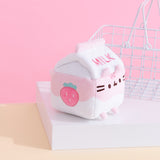 A side profile of the Strawberry Milk Sips Plush shows off the pink strawberry embroidered on the side of the plush. The plush is seated on a white pedestal with a small white wire basket in the background.  