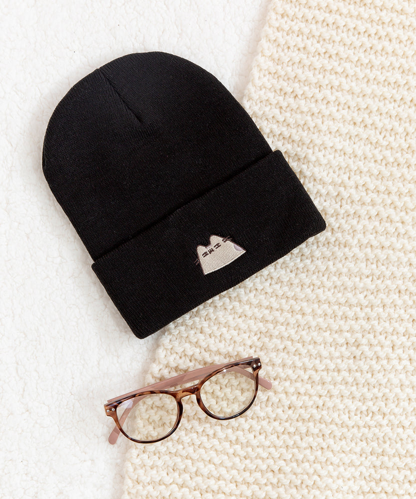 Black beanie hat laid on a white carpet surface accompanied by a cream blanket and a pair of eyeglasses.