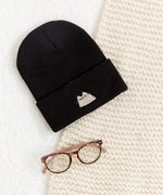 Black beanie hat laid on a white carpet surface accompanied by a cream blanket and a pair of eyeglasses.