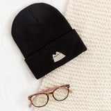 Black beanie hat laid on a white carpet surface accompanied by a cream blanket and a pair of eyeglasses.