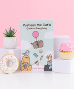 Pusheen the Cat's Guide to Everything paperback book. Blue book features Pusheen, Bo the Parakeet, Sloth, Cheek the Hamster, Little Sister Stormy, and Little Brother Pip characters on the cover artwork. The propped up book is surrounded by three white cubes with a succulent, white glazed donut, and pink cupcake.