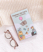 Pusheen the Cat's Guide to Everything book resting at an angle on top of a cream and white background. The book sits next to a pair of reading glasses.