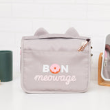 Back view of the grey toiletry bag. The back has a storage slot. Below the storage slot is a saying “Bon Meowage” in white and pink print. The “O” in Bon is a two-toned pink glazed donut.  
