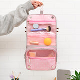 Model holds the toiletry bag open. The bag features a top hook for hanging. The interior of the tri-fold bag is light pink pola dot print and has zip-close and mesh storage compartments. The bag closes via magnets at the interior bottom of the bag. 