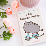 The I Am Pusheen the Cat Book on top of a white surface, with pink peonies and a pink ribbed pot around it.