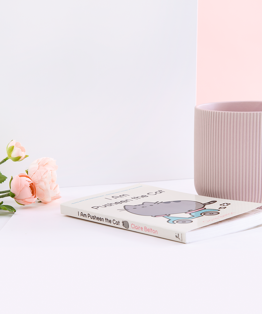 The book laying on it’s back, with pink peonies and a white wall on the left and a ribbed mug and a white wall on the right. The spine of the book is the same light yellow as the cover, with the title in brown, the author’s name in pink, an illustration of Pusheen wearing sunglasses in between the two, and the publisher’s logo on the bottom.