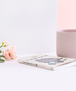 The book laying on it’s back, with pink peonies and a white wall on the left and a ribbed mug and a white wall on the right. The spine of the book is the same light yellow as the cover, with the title in brown, the author’s name in pink, an illustration of Pusheen wearing sunglasses in between the two, and the publisher’s logo on the bottom.
