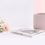 The book laying on it’s back, with pink peonies and a white wall on the left and a ribbed mug and a white wall on the right. The spine of the book is the same light yellow as the cover, with the title in brown, the author’s name in pink, an illustration of Pusheen wearing sunglasses in between the two, and the publisher’s logo on the bottom.