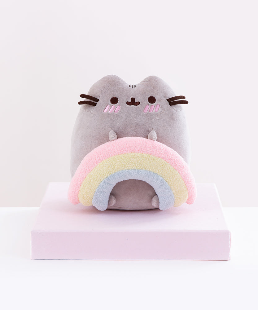 Front view of the Pusheen with Rainbow Plush sitting on top of a pink square pedestal in front of a white background. The ends of the rainbow are rounded out and vary in size, as if the rainbow is three strings of clay placed on top of one another. Pusheen’s bottom two feet peek out from underneath the inner edges of the rainbow.