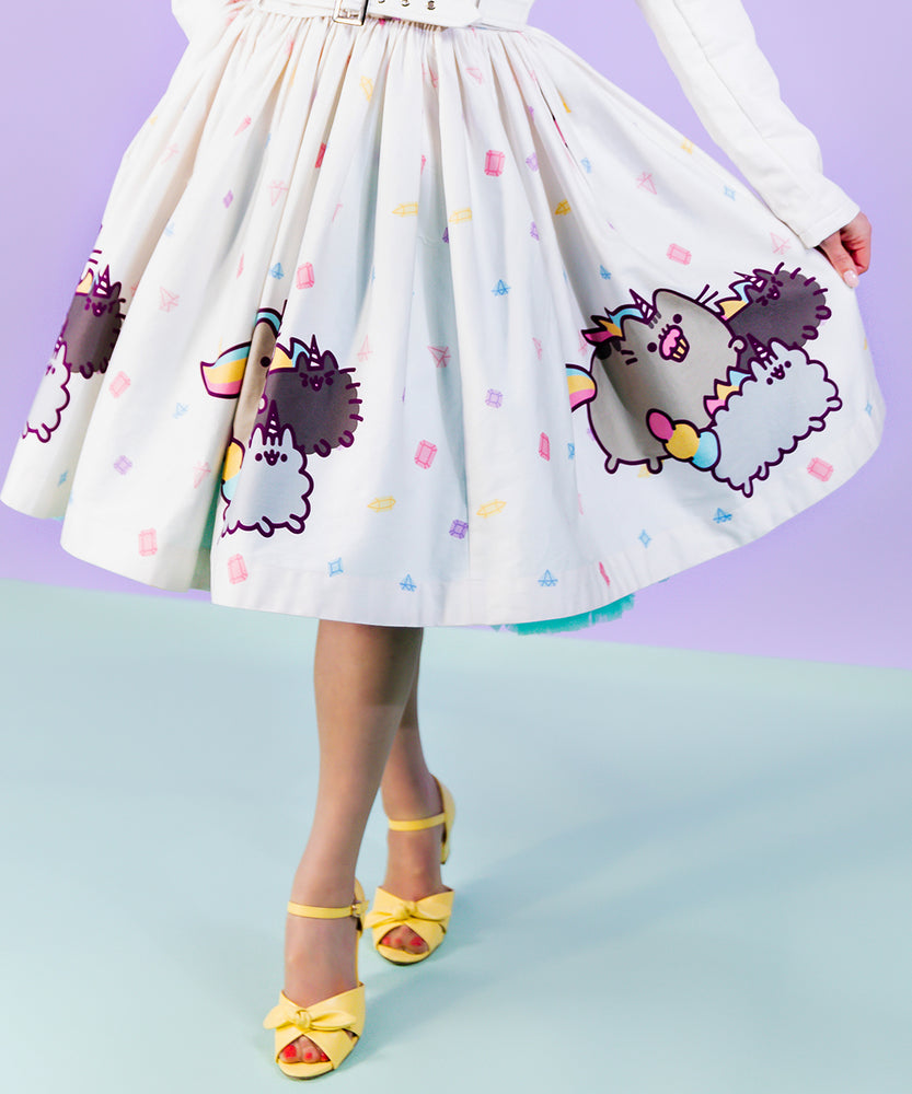 Closeup view if printed Pusheenicorn pattern skirt pattern. White skirt is dotted with pastel pink, purple, blue and yellow magical jewels with a repeating border pattern of Pusheenicorn carrying a pink cupcake in her mouth while walking alongside her and friends. 