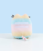 Back view of the Rainbow Pusheen Plush in front of a light blue background. Pusheen's two back stripes are white and embroidered into the plush. Pusheen's tail is a light pastel pink with white stripes. The plush is in front of a light blue background.