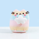 Pusheen colored in a pastel gradient, alternating between pink, yellow and blue. Pusheen is sitting upright, holding onto a plush white cloud in the shape of a star. Pusheen's stripes, eyes, and a round pink plush by her whiskers are embroidered into the plush. The whiskers are made of dark brown felt and point outwards.