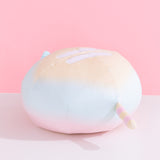 The back quarter view of the Rainbow Ombre Round Squisheen Plush sitting on top a white surface. Pusheen the Cat’s tail is striped light yellow and pastel pink striped. 