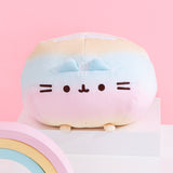 Front view of the Rainbow Round Pusheen Squisheen sitting on top of a white pedestal. A pink, yellow, and blue pastel rainbow sits in front of the plush. The yellow, blue, and pink pastel plush sits on four paws, two of which can be seen in this view.  