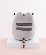 Back view of the Medium Sitting Squisheen, placed on top a square white pedestal in front of a light pink and white background. Two dark grey stripes are embroidered in Pusheen’s back, and a dark brown and light grey striped tail points out from the bottom center. Pusheen’s felt whiskers poke out from the front and are visible from the back. 