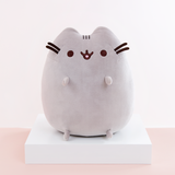 Pusheen sitting upright, with two nub paws up in the air and two nub paws at the bottom facing forward. Pusheen’s eyes and slightly open mouth are embroidered onto her face, and she has two felt whiskers poking out from each side of her face.  The medium sitting squisheen is on top of a white square podium, on top of a peach floor in front of a white background.