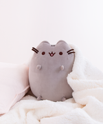 Top view of the Medium Sitting Squisheen lying decadently on top of a fuzzy white blanket, head poking in from the bottom right to lie upon a pillow. Pink peonies are resting on top of the pillow as well in the upper left corner, right above Pusheen.