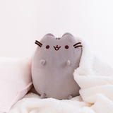 The Medium Sitting Squisheen Plush cozily seated besides a white pillow, with a white fuzzy blanket draped over her.