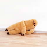 Sloth lounging on his side, right hand propping up his head and his left arm draped over his chest. Sloth is laid upon a wooden floor in front of a white background. He’s a real charmer, this lad.