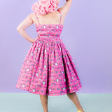Back view of the dreamy pink dress shows off the pleating at the top of the skirt. Model is wearing yellow shoes and a pink wig while standing with one hand on the hip and one hand on the head. 