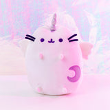 The Super Pusheencorn plush in front of a magical fluorescent background, bathed in a magenta light. The Super Pusheenicorn plush reflects the light around her and appears light pink as a result, her Moon mark appearing to be a darker purple.