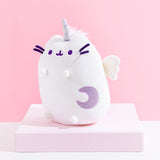 Quarter view of the Super Pusheenicorn Plush facing the left on top of a white square pedestal, in front of a pink background. The sparkly purple crescent moon on Pusheen’s front and the plush wing is more visible from this angle, though the multicolor mane is only visible directly behind Super Pusheenicorn’s horn.