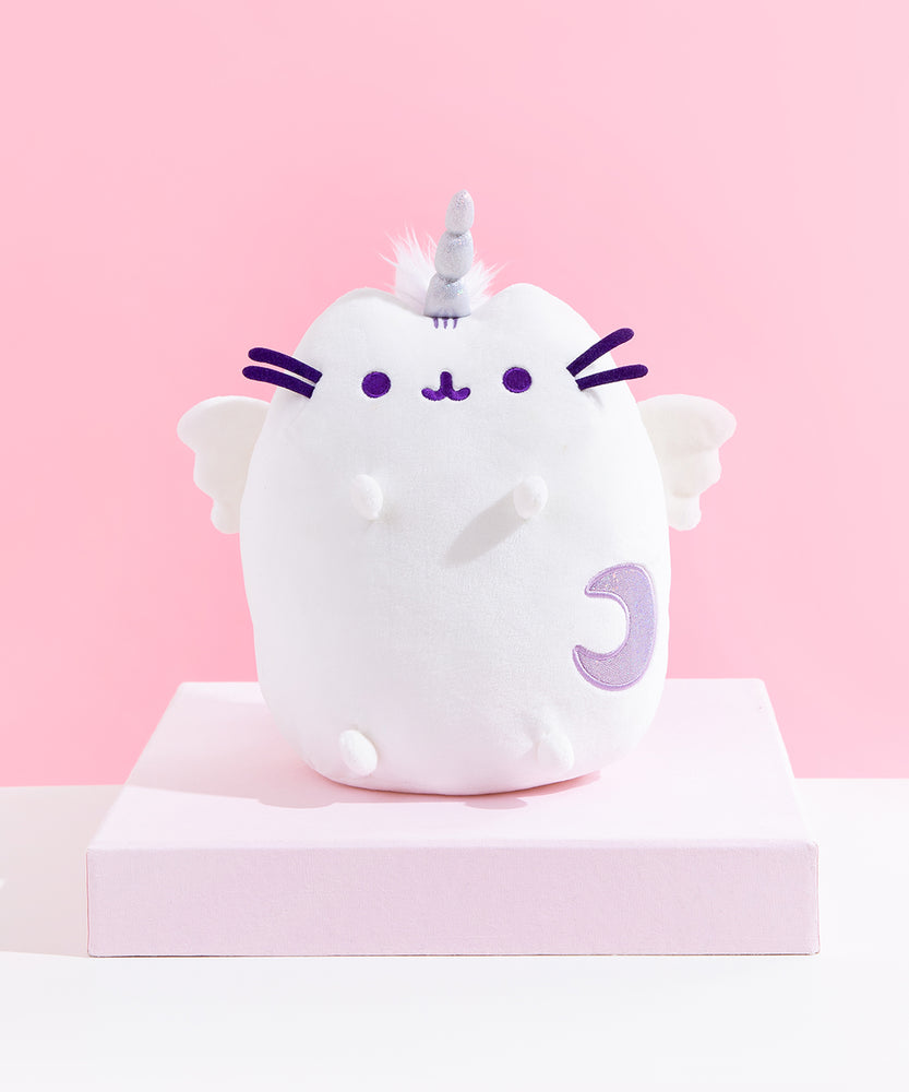 The Super Pusheenicorn Plush on top of a white square pedestal, in front of a pink background. Little tufts of white fur is visible behind Super Pusheenicorn’s horn.
