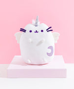 The Super Pusheenicorn Plush on top of a white square pedestal, in front of a pink background. Little tufts of white fur is visible behind Super Pusheenicorn’s horn.