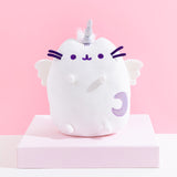 The Super Pusheenicorn Plush on top of a white square pedestal, in front of a pink background. Little tufts of white fur is visible behind Super Pusheenicorn’s horn.