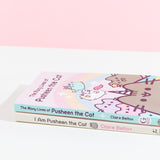 The Many Lives of Pusheen the Cat stacked on top of the previous Pusheen book, I Am Pusheen the Cat. Both books are paperbacks of similar length, though the spine for Many lives is mint colored with purple font and a white sparkle, while the first book is a light yellow with brown and pink text with a sunglasses wearing Pusheen on the spine.