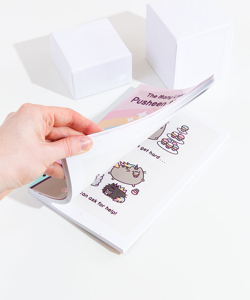 A model’s hand partially opening the book, showing a preview of a comic featuring Puseenicorn and family with cupcakes. 