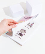 A model’s hand partially opening the book, showing a preview of a comic featuring Puseenicorn and family with cupcakes. 