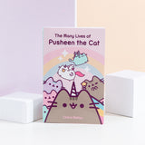 A paperback book being propped up by two white blocks in front of a pink and yellow background. The book is titled ‘The Many Lives of Pusheen the Cat’ and features Pusheenicorn in the middle, a winking Mermaid Pusheen on the right, the Pastel Pusheen on the left, Super Pusheenicorn carry a cookie and Dragonsheen flying above in front of a rainbow with sparkles. 