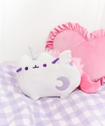 The Super Musical Pusheenicorn Plush nestled on top of a purple and white plaid blanket, pink heart pillow outlined with ruffles behind her.