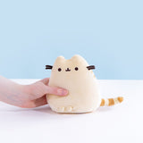 A model’s hand holding the Yellow Mini Squisheen Plush in front of a white and blue yellow background. The yellow plush has dark yellow stripes that almost appear orange.