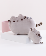 The Medium Pusheen Plush on a white square platform, with a mini Pusheen plush angled in front as a size comparison for the Medium Plush. The two plush are in front of a pink ribbed pot and a light grey background.