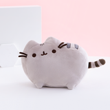 The Medium Pusheen Plush on top of a white floor, in front of a pink background and two white shapes, a big square and a small hexagon. The Pusheen Plush features a grey cat with a few dark stripes standing in profile. The front and back left legs are visible from the front, both ears, her felt whiskers, and her embroidered face are faced directly to the front, and a small striped tail sticks out immediately from the side. The plush is very rounded out, with no hard edges.