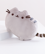 The Medium Pusheen plush on top of a white square platform. A pink peony is visible on the left in the background, and the plush is in front of a light pink background.